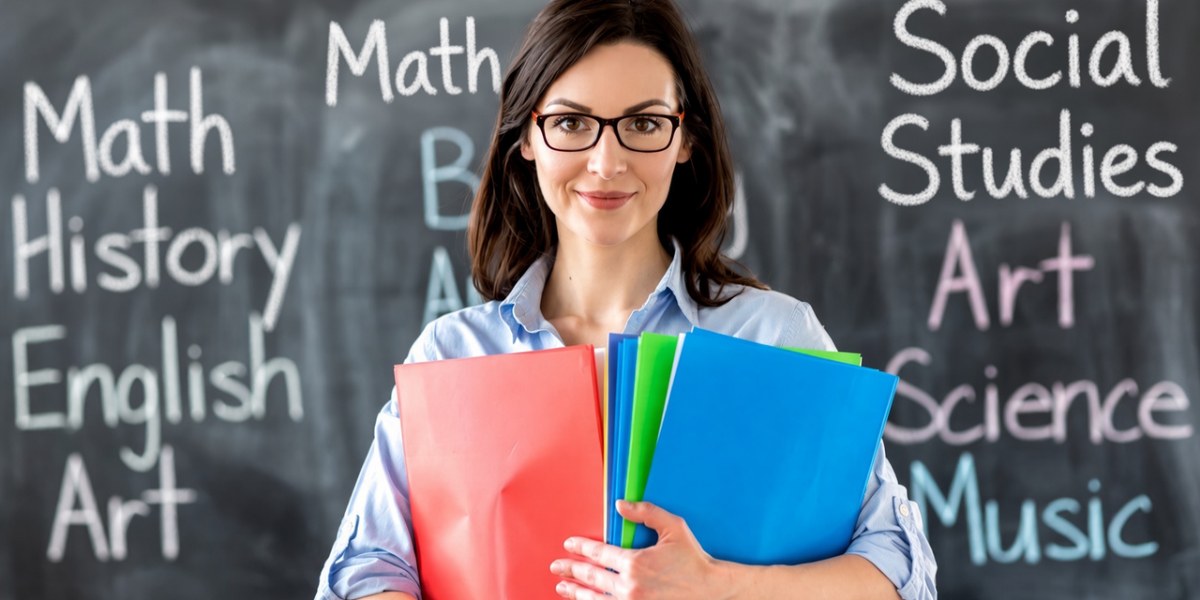 A teacher holding folders in front of a chalkboard listing school subjects.